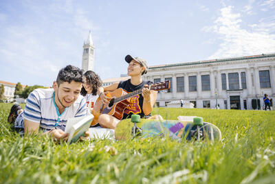 Three students on Memorial Glade, reading and playing guitar